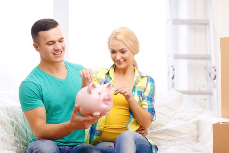 A young couple is smiling while holding a piggy bank and surrounded by boxes in the concept of 'how to save money when moving to Croydon'.